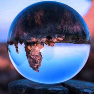 A clear glass sphere on a tree stump reflects and inverts a rocky landscape with trees under a blue sky at sunset, capturing the essence of Earth and Space Science AB in one captivating image.