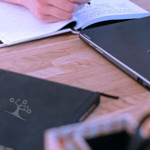 A person writes in a notebook by an open laptop on a wooden desk, surrounded by English Language Arts, Grade 10 AB materials—a closed notebook, pen holder, and scissors nearby.