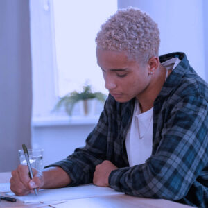 A person with short, curly blond hair sits at a table working on English Language Arts, Grade 12 AB. Wearing a plaid shirt over a white T-shirt, they write on paper with a pen. A glass of water and calculator are nearby; curtains and a plant are behind them.