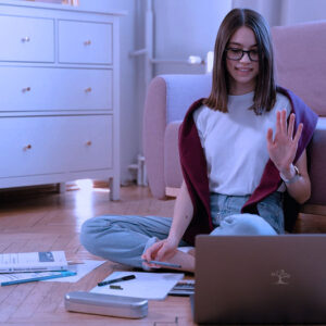 A young woman sits cross-legged on the floor in her cozy room, waving at her laptop as she studies English Language Arts, Grade 9 AB, surrounded by papers and stationery.
