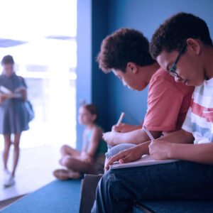 Two boys sit and write in notebooks on a bench, focused on their work. In the blurred background, a girl stands reading and another girl sits on the floor. A stylized tree logo is visible on the wall.