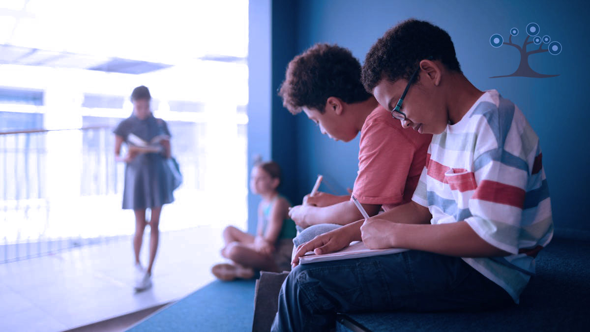 Two boys in the foreground writing in notebooks and two girls in the background out of focus with one of the girls reading a book.