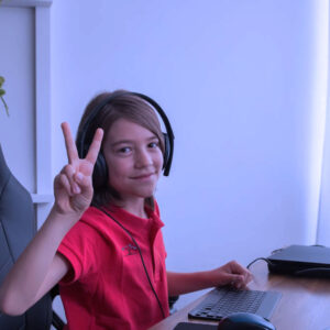 A smiling child wearing a headset sits at a computer desk and flashes a peace sign with their fingers. The room is softly lit with natural light from a window and a plant hangs nearby.