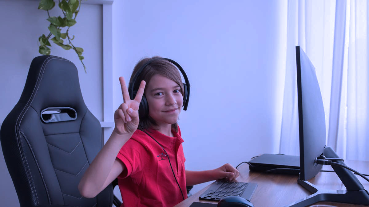 A student sitting at a desk in front of a computer while holding up two fingers similar to the sign for peace.