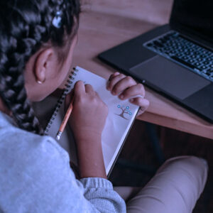 A child with braided hair sits at a desk, drawing in a notebook with a pencil. An open laptop is on the desk beside them. The scene suggests studying or creative work at home.