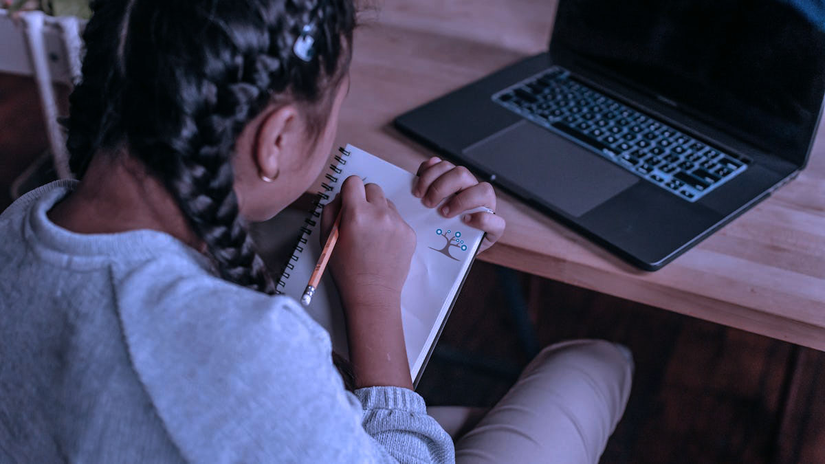 A girl with a pencil in her hand writing in a notebook while sitting a desk with a laptop on it.