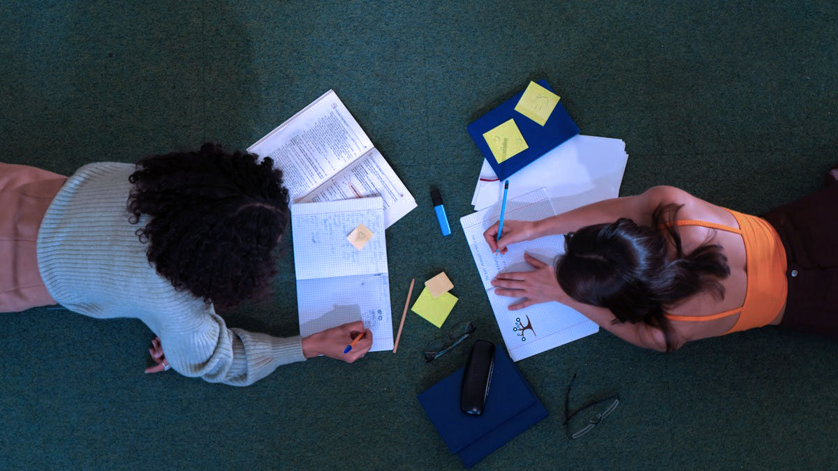 An overhead shot of two girls laying on the floor doing homework.