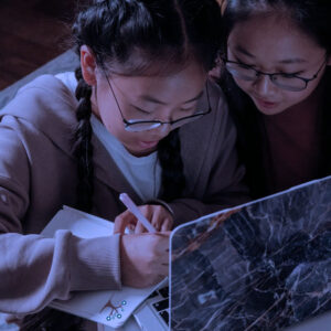 Two girls with glasses sit closely together on the floor, working on a laptop. One writes in a notebook while the other looks at the screen. The scene is warmly lit and they appear to be focused.