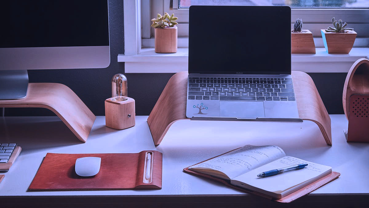 A desk with lapton on a stand and a open notebook and other desk items.