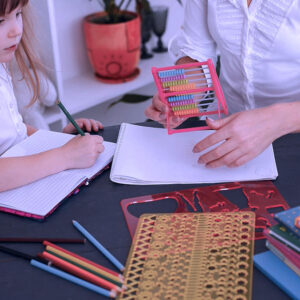 A young girl writes in a notebook at a table while an adult beside her holds an abacus. Colored pencils, notebooks, and drawing stencils are scattered on the table.