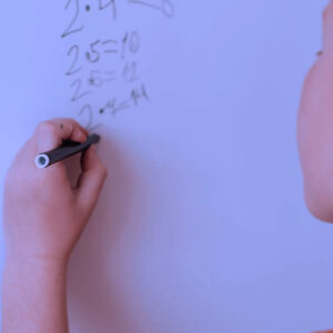 A child writes math equations with a marker on a whiteboard. The child is facing away from the camera, and part of their face and hand are visible. There is a small tree logo in the bottom left corner.