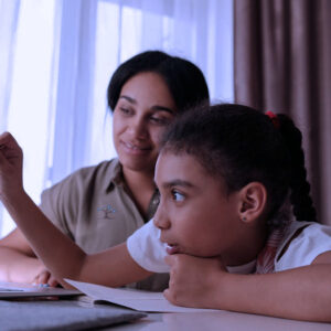 A woman helps a young girl with her homework, pointing at a laptop screen with a pencil, while the girl watches attentively, with an open notebook in front of her.