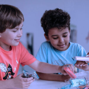 Two kids sitting at a tabel with bags in their hands whicl ea teacher is handing them additional bags.