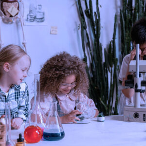 Three children using Science, Grade 3 work together at a table—one looks through a microscope, another examines a sample, while the third smiles. Science lab equipment and plants are arranged around them.