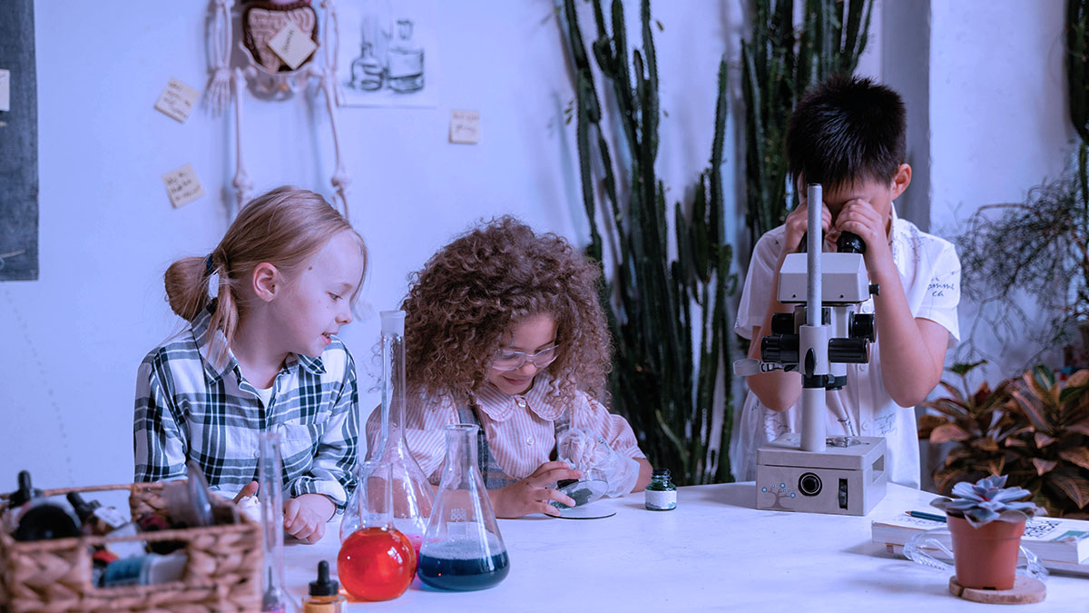 Three elementary students in a science room with one of them looking through a microscope.