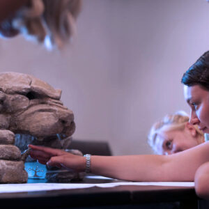 A student building a stone cave model while other students look on.