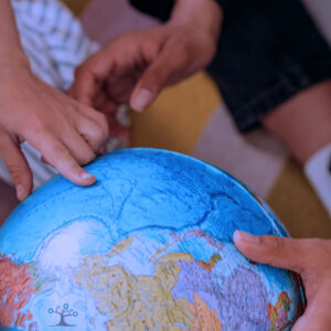 Three people sit together, closely examining and pointing at locations on a blue and colorful globe. Their hands rest on the globe, indicating curiosity and learning about geography.