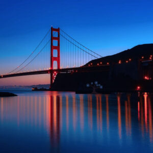 The Golden Gate Bridge at dusk, illuminated with red lights, spans across calm blue water, with a dark hill silhouette and a clear twilight sky in the background.