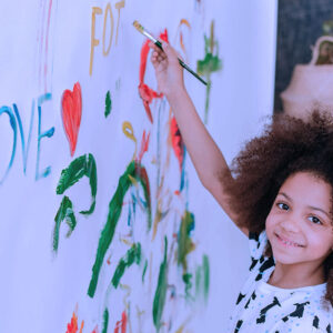 A smiling child with curly hair paints colorful words and shapes, including LOVE and a red heart, on a large white wall. The child looks at the camera, holding a paintbrush.