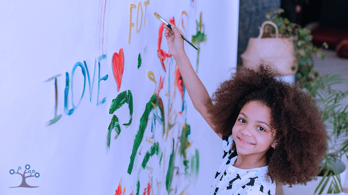 Girl smiling while standing at a white wall with a painting brush in her hand.