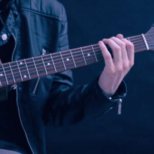 A person in a black leather jacket plays an acoustic guitar, focusing on their left hand on the fretboard—capturing the diverse influences explored in Ethnic Studies. The background is dark and blurred.