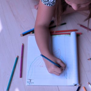 A child sits on a light wooden floor, drawing creative designs in a spiral notebook with the Art 2-3 colored pencil set scattered around, holding a blue pencil from Art 2-3 to sketch on the paper.