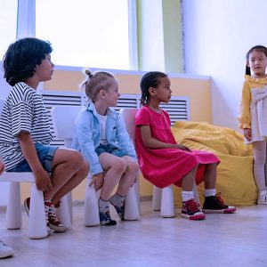 In a brightly lit classroom, a young girl in a yellow dress points to numbers on the Math, Grade K-1 wall chart as four children watch her from a bench.